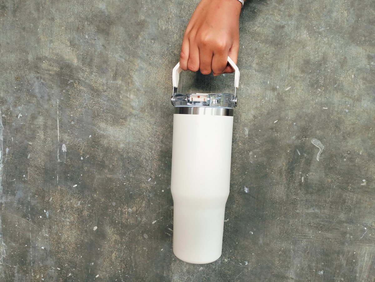 Close up photo of a hand holding a white tumbler against a grey concrete wall background. Minimalist drinkware and reusable container concept with copy space, suitable for branding and mockup purposes.